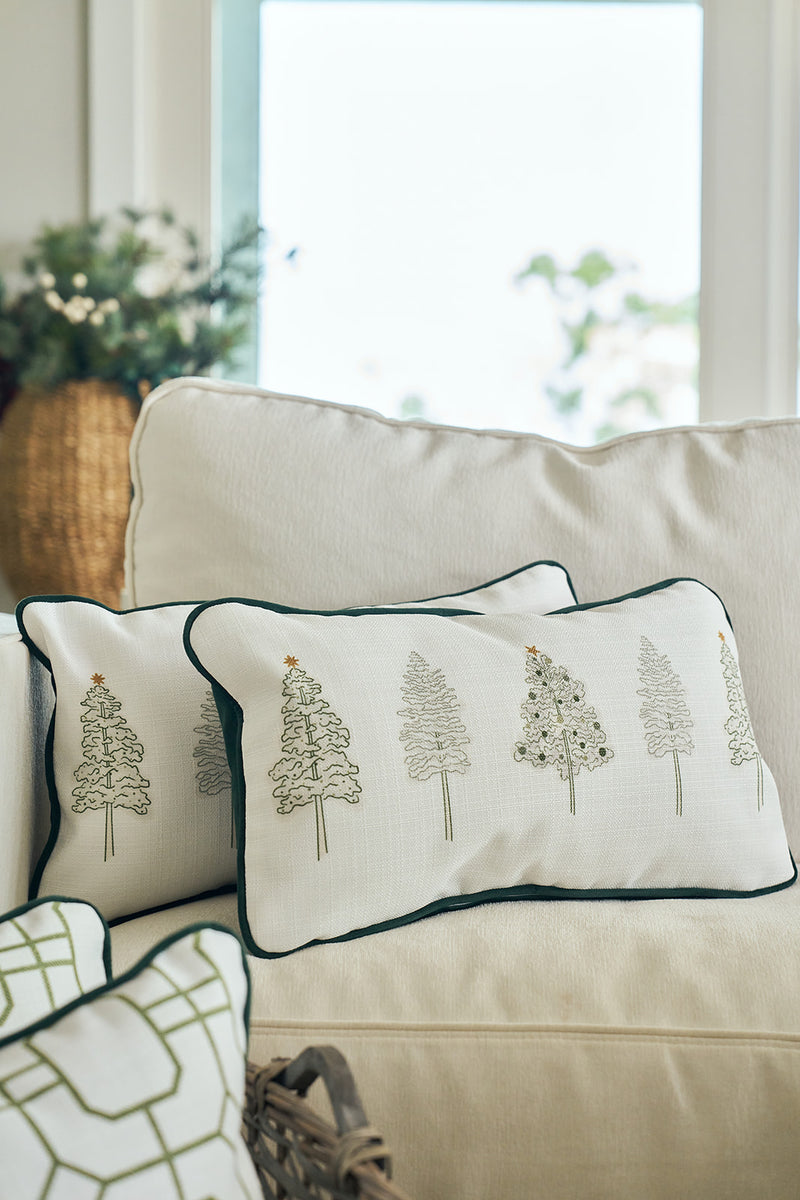 The Row of Trees Extended Lumbar Pillow with embroidered trees rests on a light beige couch, with a woven basket filled with greenery in the background as sunlight streams through the window, creating a festive holiday scene.