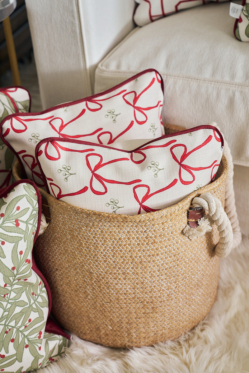 A woven basket with rope handles holds two white pillows featuring red ribbon, green bows, and mistletoe designs, placed beside a cream-colored couch styled with Bows & Mistletoe Lumbar Pillows.