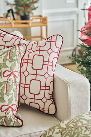 Two decorative pillows rest on a beige couch: one is the Red Holiday Lattice Oversized Pillow, while the other features green mistletoe with a red bow. In the background, a small Christmas tree and festive holiday decor are visible.