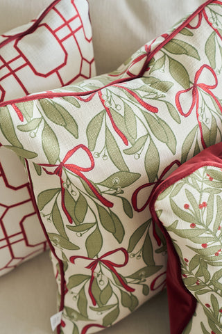 Three holiday pillows with red and green patterns rest on a light surface. Two have leafy green and red ribbon designs, while the third is the Oh Mistletoe Oversized Pillow with a geometric red and white mistletoe pattern.
