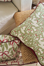 A Jolly Holly Pillow and a fabric basket with green leafy and red berry patterns are arranged on a woven rattan seat next to a white upholstered chair.