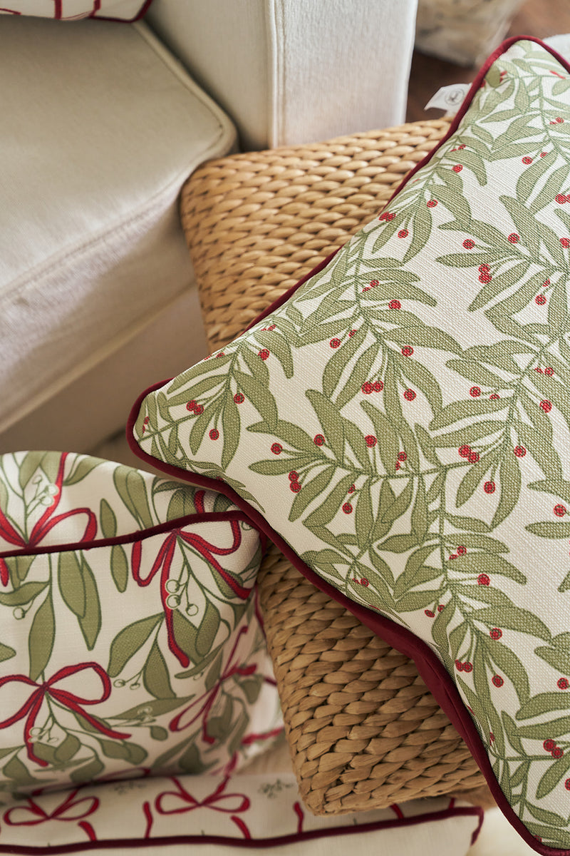 A Jolly Holly Pillow and a fabric basket with green leafy and red berry patterns are arranged on a woven rattan seat next to a white upholstered chair.