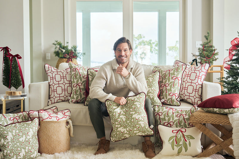 A man in a cozy sweater relaxes on a white sofa, leaning against the Jolly Holly Pillow. Festive holiday pillows and small Christmas trees surround him, filling the bright living room with warmth and cheer.