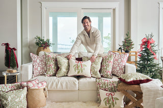 A man in a white sweater places the Oh Mistletoe Oversized Pillow on a white sofa in a holiday-themed living room with small Christmas trees, red ribbons, and big windows.