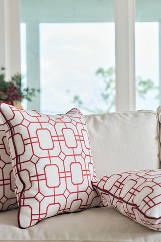 Two white throw pillows, including a Red Holiday Lattice Oversized Pillow, rest on a cream-colored sofa in front of large, bright windows with a blurred outdoor view in the background.