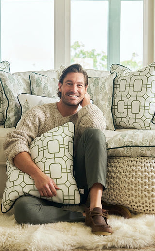 A man sits on a rug, smiling and holding the Green Holiday Lattice Oversized Pillow from Sewing Down South, with several matching holiday decor pillows displayed on the sofa behind him.