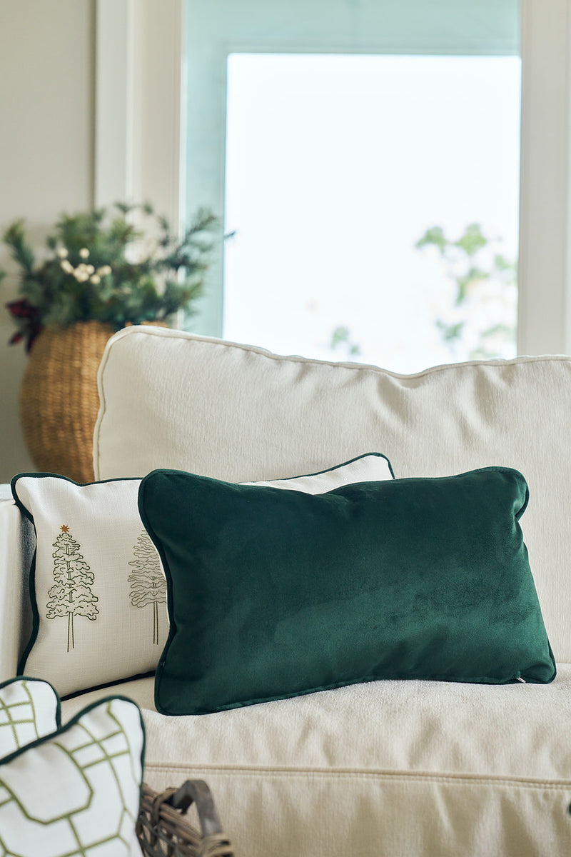 A green velvet holiday pillow sits on a cream couch next to the Row of Trees Extended Lumbar Pillow, which is white and embroidered with green trees. In the background, a woven basket with greenery rests by a sunlit window.