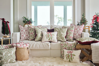 A white couch is decorated with festive pillows, including the Red Holiday Lattice Oversized Pillow with green, white, and red patterns. More oversized holiday pillows and small decorated Christmas trees add cheer to the sunlit, cozy room.