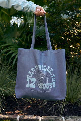 A person holds the SDS Nashville 12 South Tote Bag with Nashville, TN South 12 and Were Awfully Glad Youre Here printed on it. This cotton canvas bag features a graphic design set against greenery and brick edging.