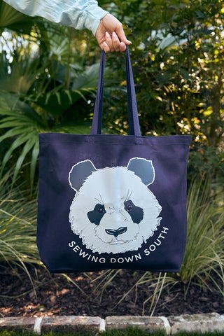 A person holds a navy blue Panda Tote Bag featuring a panda face and Sewing Down South text, with a background resembling a UFO landing site surrounded by lush green foliage, suggesting an outdoor setting.