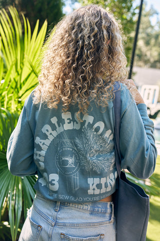 Someone with curly hair stands outdoors in a blue SDS King Street Long Sleeve Tee featuring Charleston and a design on the back. Theyre holding a blue tote bag, walking by the iconic Charleston Storefront, surrounded by lush greenery and sunlight.