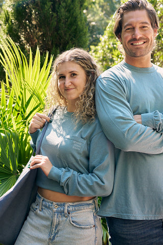 A curly-haired woman and a bearded man, both in long-sleeved blue shirts, are smiling by a pool amidst lush greenery. The woman confidently carries her chic SDS Nashville 12 South Tote Bag, posing outdoors on this sunny day.