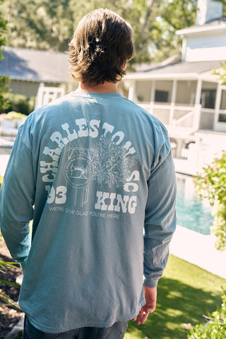 A long-haired person stands by a pool wearing a blue SDS King Street Long Sleeve Tee with palm trees, displaying Charleston, SC and Were Sew Glad Youre Here, from the bustling Charleston Storefront. Greenery and a house are in the background.