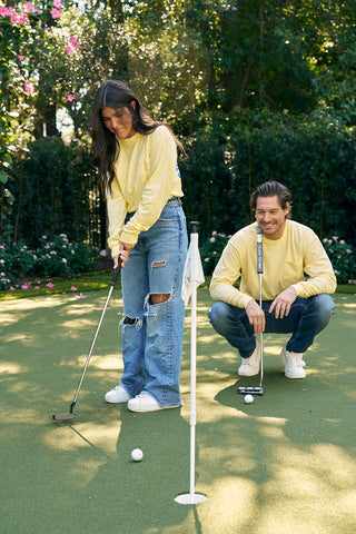 On a sunny day near the Charleston Storefront on King Street, a woman wearing SDS King Street Long Sleeve Tee and ripped jeans putts a golf ball on a small green. A man in the same tee crouches nearby, surrounded by trees and flowers.