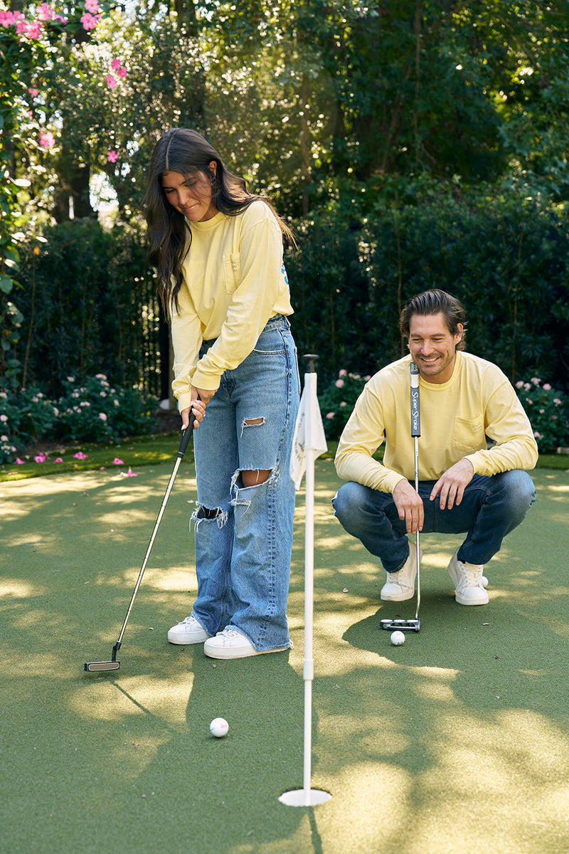 On a sunny day near the Charleston Storefront on King Street, a woman wearing SDS King Street Long Sleeve Tee and ripped jeans putts a golf ball on a small green. A man in the same tee crouches nearby, surrounded by trees and flowers.