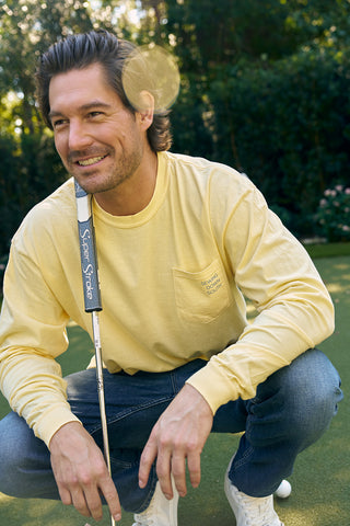 A man with medium-length hair in an SDS King Street long sleeve tee and jeans squats on the King Street putting green, holding a golf club. Sunlight filters through the trees, casting a warm glow reminiscent of Charlestons charming storefronts.