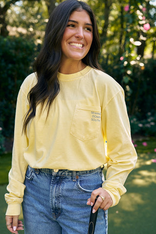 A person with long dark hair smiles while holding a mini-golf club, wearing a yellow SDS King Street Long Sleeve Tee and blue jeans, standing amid greenery and pink flowers. The vibe matches the vibrancy of the SDS Charleston Storefront on King Street.