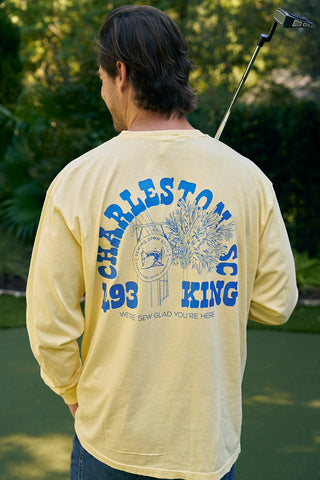 A person in a yellow SDS King Street Long Sleeve Tee with a blue tree graphic and Charleston, SC 843. Were sew glad youre here across the front, stands on a lush green lawn holding a golf club over their shoulder, embodying the vibrant energy of King Street.