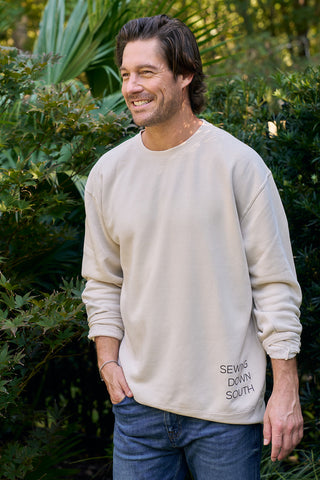 A brown-haired, light-bearded man smiles outdoors before lush greenery, confidently wearing a SDS ‘King Street’ Crewneck Sweatshirt in Bone with blue jeans, as if about to visit the Charleston Storefront.