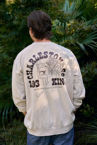 A man stands outdoors facing away, wearing the SDS King Street Crewneck Sweatshirt in Bone. The back features CHARLESTON CO. 193 KIN, plant and sun graphics, and WERE SEW GLAD YOURE HERE beneath the design.