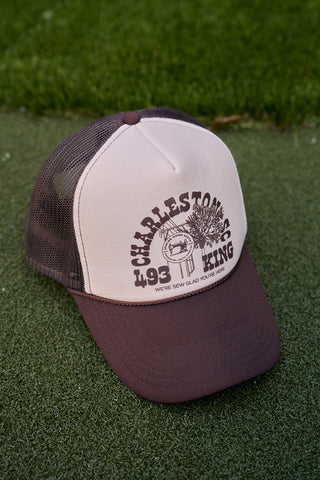 A brown and beige SDS King Street Trucker Hat sits on grass, showcasing Charleston SC 1995 King with a pineapple and palmetto tree illustration for Charleston Storefront charm. Below it reads Were Sew Glad Youre Here.