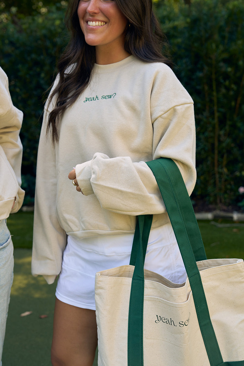 A smiling person outdoors, surrounded by lush greenery, wears a beige sweatshirt with yeah, sew? text and holds a Yeah, Sew? Canvas Tote Bag—beige with green straps and the same playful phrase.