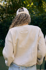 Amidst lush greenery, the curly-haired person sports a beige sweatshirt and backwards cap. Their look features the Yeah, Sew? Embroidered Cropped Sweatshirt from the Fall/Winter cozy collection, made of recycled fleece for ultimate comfort and style.