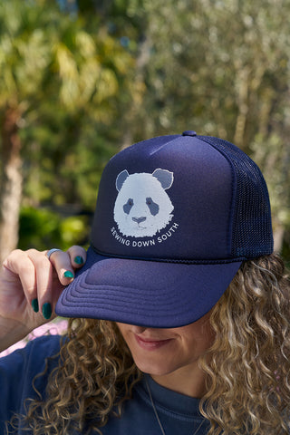 A person with curly hair wears the Panda Trucker Hat, a navy blue cap with a white panda face and embroidered SDS collegiate logo, perfect for Panda People. Outdoors among trees, they show green nails while holding the bill.