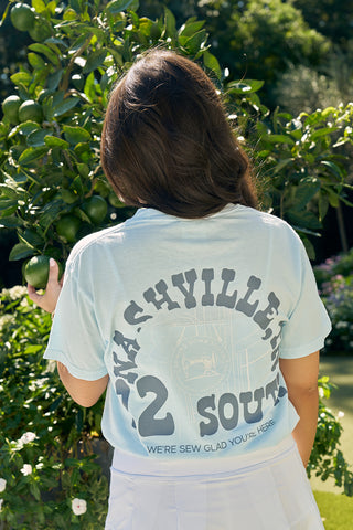 A person with long brown hair stands outdoors, picking green fruit from a tree while wearing a light blue SDS Nashville '12 South' Tee featuring "Nashville 12 South" and "We’re sew glad you’re here" printed on the back.