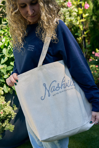 A curly blonde-haired person in a navy sweater holds an SDS Nashville Rope Tote Bag made of heavy cotton canvas while standing in a garden adorned with green plants and pink flowers.