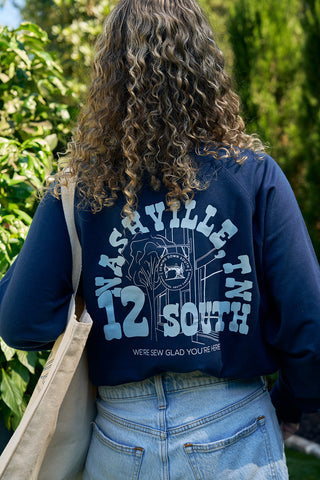 Outdoors, a person with curly hair wears an SDS Nashville 12 South Crewneck Sweatshirt in cozy navy, paired with denim shorts and a canvas tote bag. The sweatshirts text, echoing Sewing Down South’s Charleston Storefront charm, reads Were sew glad youre here.