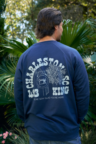A person with dark hair stands outdoors, facing away in an SDS King Street Crewneck Sweatshirt in indigo, featuring white graphics and text including Charleston and a palm tree, set against lush green plants.