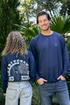 Two casually dressed people outdoors: the left person has curly hair, wearing a navy sweatshirt with Charleston King from the King Street Crew collection. The smiling person on the right wears an Indigo SDS King Street Crewneck Sweatshirt with Goodbye Good Vibes on the front.