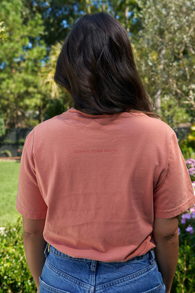 A person with long dark hair stands outdoors in a green garden setting, wearing jeans and a coral SDS Nashville Rope T-Shirt that features SEWING DOWN SOUTH on the back.