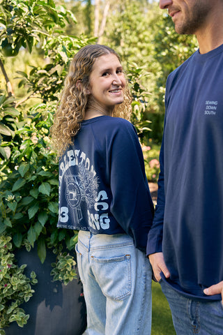 A curly-haired woman smiles in an SDS King Street Crewneck Sweatshirt, Indigo, featuring a back logo. Beside her, a man in a matching blue shirt is partially visible. Theyre outdoors amid lush greenery, echoing the vibrant feel of the King Street Crews Charleston storefront.
