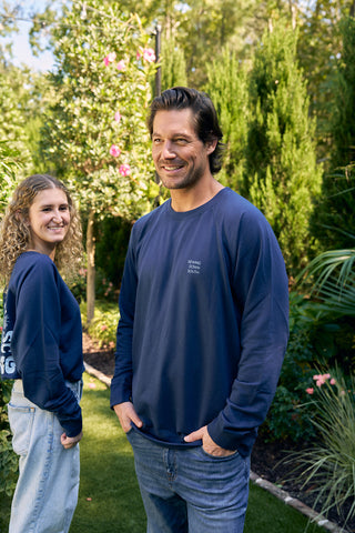 Two people in a garden, both wearing SDS King Street Crewneck Sweatshirts in Indigo. The man stands and smiles, while the woman turns slightly with a grin. Amidst lush greenery and sunshine, they resemble members of the King Street Crew enjoying downtime.