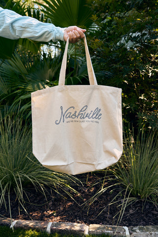 A person holds a beige SDS Nashville Rope Tote Bag against green plants. Made from heavy cotton canvas, it displays Nashville in large script with were sew glad youre here below, making it an ideal travel keepsake.