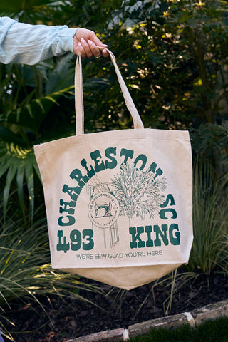 Someone holds an SDS King Street Tote Bag made of 100% cotton canvas, displaying green Charleston SC, 193 King, Were Sew Glad Youre Here text with plant and water tower illustrations; lush greenery is in the background.