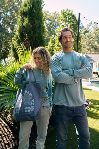 A woman and a man stand outdoors on a sunny day by the Charleston Storefront, both wearing SDS King Street Long Sleeve Tees in light blue. The woman holds a large blue tote bag, surrounded by greenery with a pool and house seen in the background.
