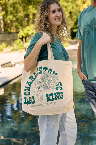 A smiling woman with curly hair carries an SDS King Street Tote Bag—100% cotton canvas, featuring green CHARLESTON SC text—over her shoulder by a pool, next to a friend. Both wear casual green shirts.