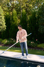 A man in a light pink SDS King Street Illustration Crewneck and jeans uses a long pool skimmer to clean a backyard pool, with green trees, bushes, and pink flowers in the background.