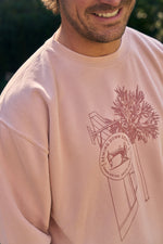 A person wears the SDS King Street Illustration Crewneck in light pink, featuring a graphic of a tree, airplane, and circular Sewing Down South, King Street, Charleston, South Carolina logo. The lower half of their smiling face is visible.