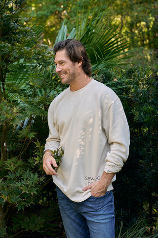 A man with medium-length brown hair and a beard, smiling and looking left, stands outdoors among green plants wearing blue jeans and the SDS King Street Crewneck Sweatshirt in Bone.