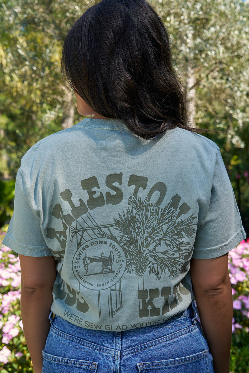 A person with long hair faces away, wearing a light green SDS King Street Flagship T-Shirt made of 100% ring-spun cotton, featuring printed text and graphics. The background is filled with trees and colorful flowers, evoking a charming Charleston storefront atmosphere.