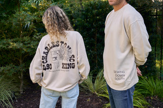 Two people outside near a Charleston storefront wear the SDS King Street Crewneck Sweatshirt in Bone; one shows the back graphic with Charleston, SC, while the other displays Sewing Down South on the front.