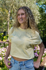 A woman with curly hair beams in a garden, wearing the vibrant yellow SDS King Street Flagship T-Shirt and blue jeans. Pink flowers and lush green foliage provide a charming backdrop, reminiscent of the Charleston Storefront.