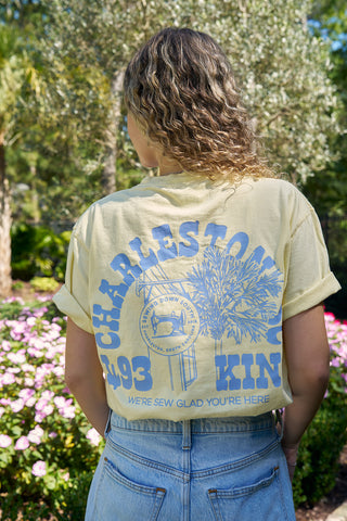 A person with curly hair, wearing a light yellow SDS 'King Street' Flagship Tee and light denim jeans, stands in a garden with pink flowers and greenery, facing away from the camera.