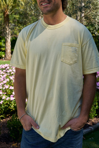 A man stands in a garden with pink flowers, smiling slightly with hands in his jeans pockets, wearing a light yellow SDS 'King Street' Flagship Tee made from heavyweight cotton.