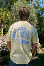 A person with brown hair stands in a garden of pink flowers and palm trees, wearing the vibrant 100% ring spun cotton SDS King Street Flagship T-Shirt. The shirt features a clock tower design, CHARLESTON, KING, and celebrates your visit to this charming Charleston storefront.