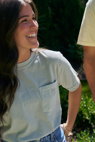 A woman with long brown hair smiles outdoors, wearing an SDS 'King Street' Flagship Tee with a pocket and blue jeans. A man in a similar shirt stands beside her, surrounded by lush greenery.
